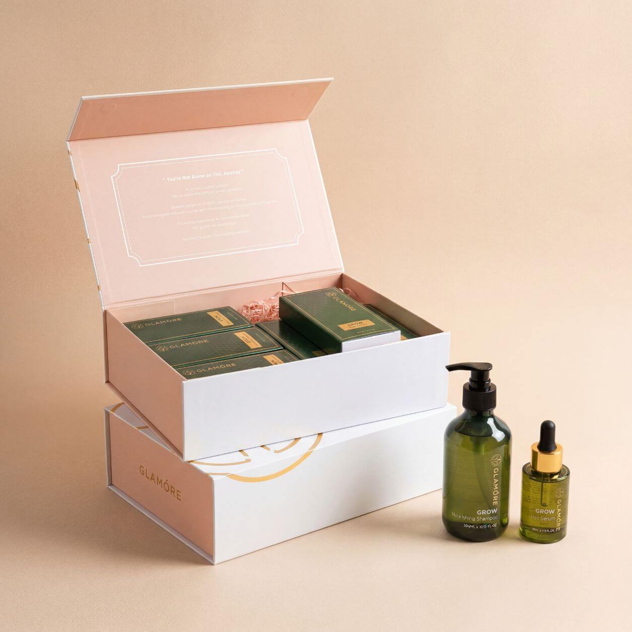 An open premium gift box with pale blush pink and white packaging, containing green boxed skincare products, shown beside two dark green cosmetic bottles with black and gold droppers on a warm beige studio background. The brand name GLAMORE is visible on the lower box and bottle labels.