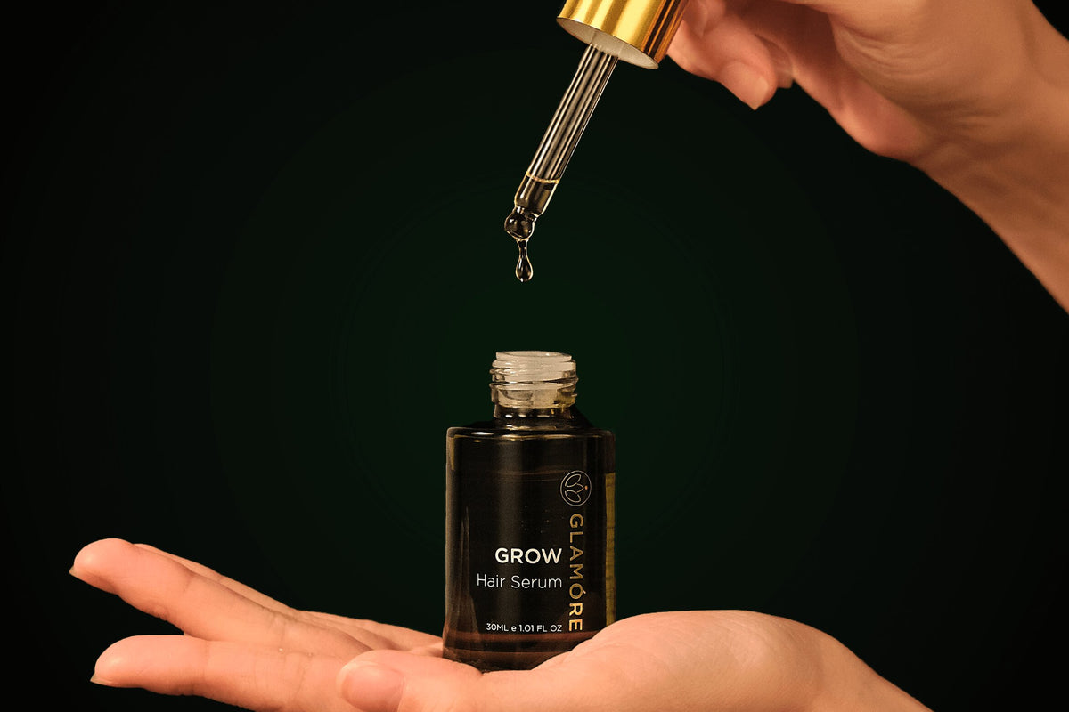 A hand holds an open bottle of Glâmeré Grow Hair Serum while another hand drops the serum from a gold-rimmed glass dropper above it, against a dark green background.
