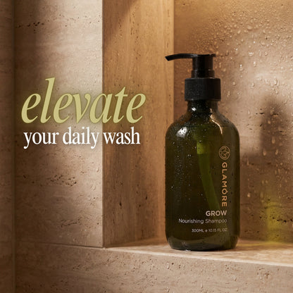A dark olive green pump bottle of GLAMORE GROW Nourishing Shampoo sits on a beige stone ledge against a matching textured wall with water droplets. The text 'elevate your daily wash' is displayed on the left side.